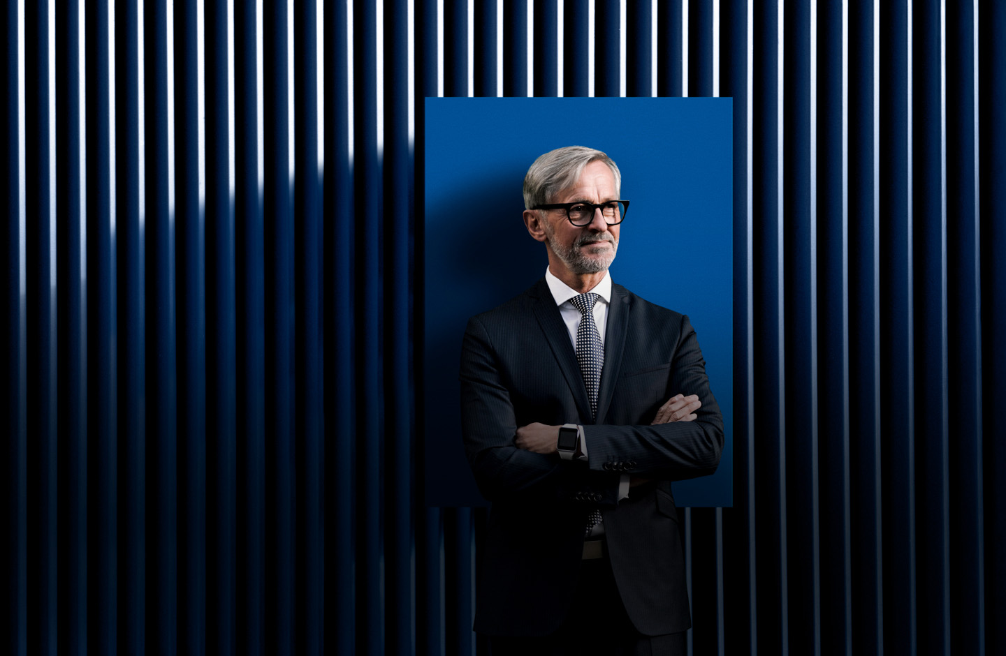 Ein älterer Mann steht mit Anzug vor einer blauen Wand.